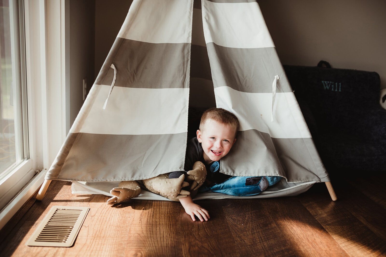 A little boy plays in his teepee while peeking out and laughing.
