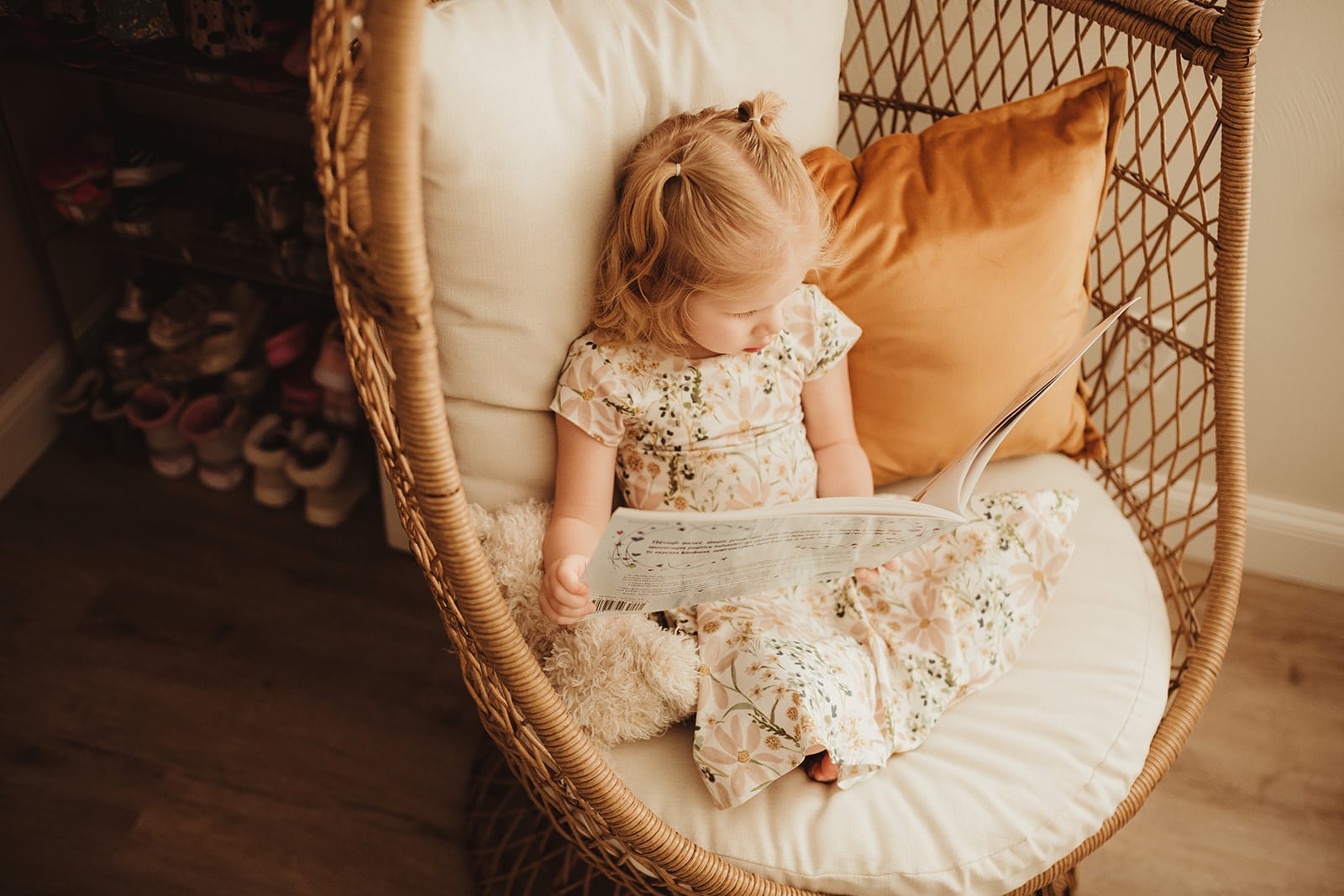 A little girl wearing a floral dress is reading a book while sitting in a boho wicker chair.